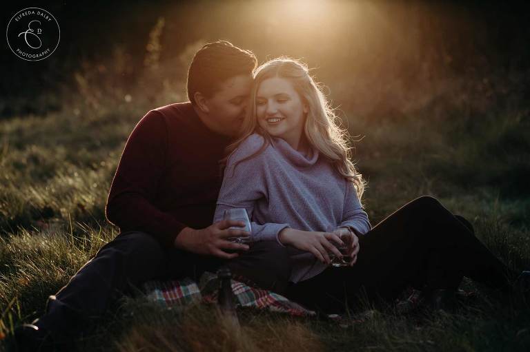 Engaged couple sitting in a field and enjoy champagne with golden light behind them