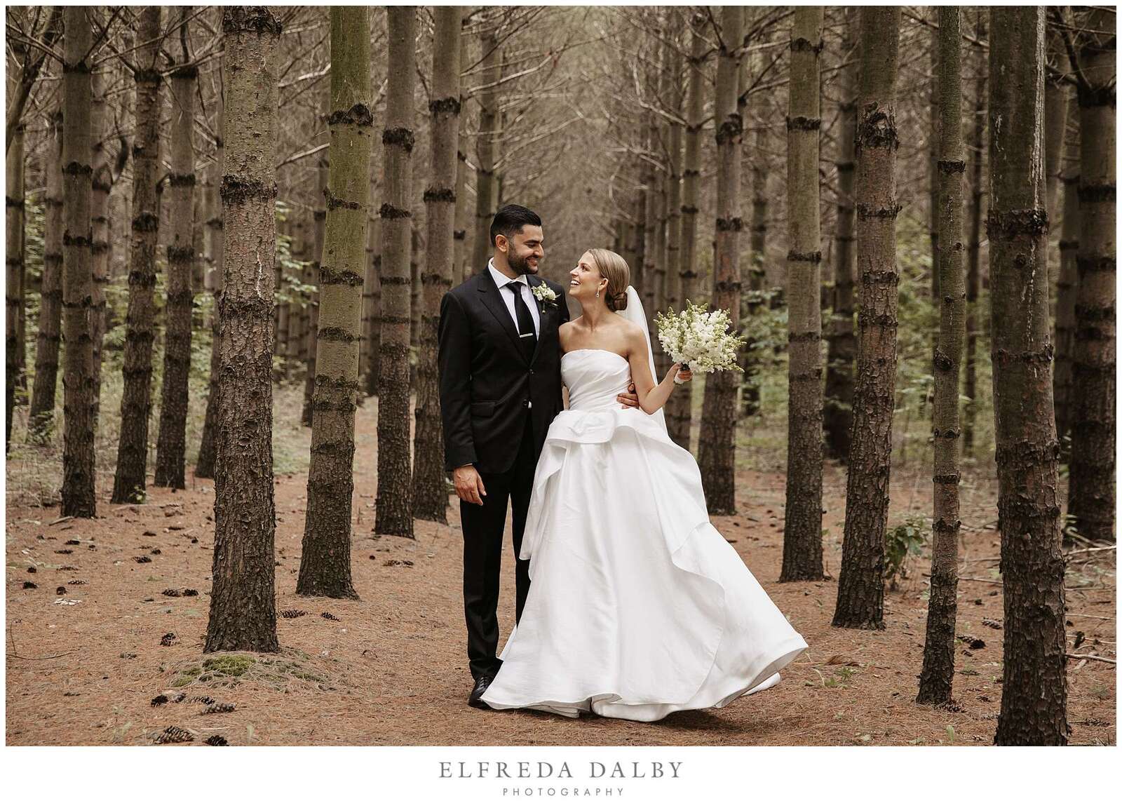 Bride and groom standing between a row of pine trees